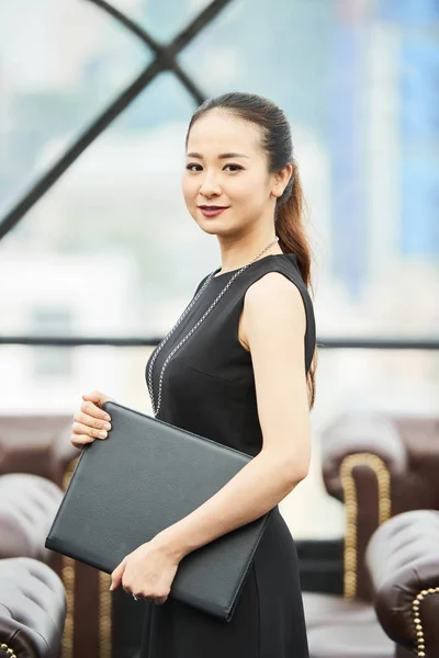 Portrait of young Asian businesswoman wearing black dress holding folder with documents and looking at camera at office