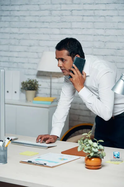 インドのオフィスワーカーは テーブルの近くに立って オフィスでコンピュータモニターを見ながら 携帯電話で会話をしています — ストック写真
