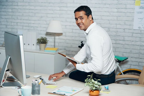 Portrait of Indian office worker sitting at the with tablet pc smiling at camera while typing on computer keyboard at office