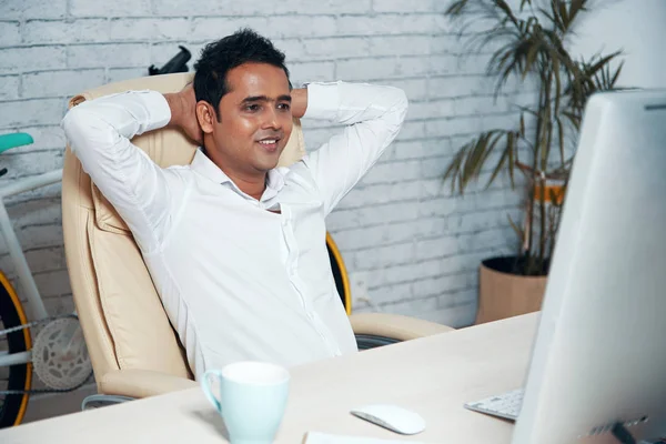 Indian smiling businessman sitting in comfortable chair in front of computer monitor and relaxing after hard work at office