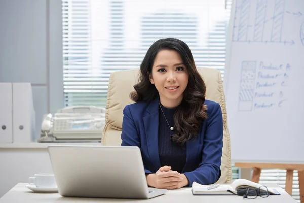 ラップトップコンピュータの前で彼女の職場に座って カメラに微笑むアジアの若いビジネスウーマンの肖像画 — ストック写真