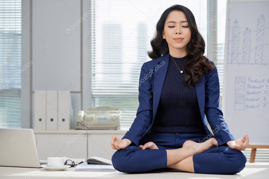 Young beautiful businesswoman sitting at the table in lotus position with her eyes closed and meditating during break at office