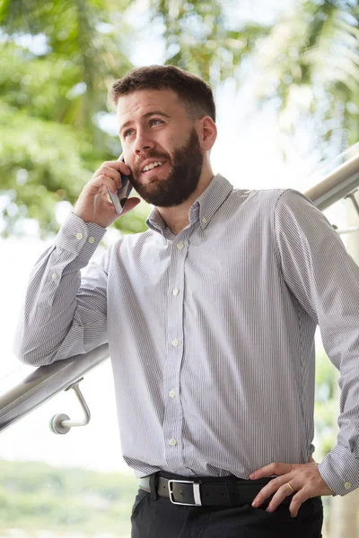 Young bearded office worker standing and listening to somebody on mobile phone while standing on staircase at office
