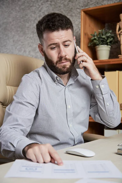 Serious bearded financial consultant sitting at the table pointing at document and consulting his client on mobile phone at office