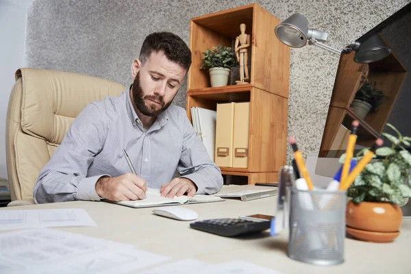 ひげを生やした若いマネージャーがオフィスデスクに座り メモ帳に新しい計画やアイデアを書いている — ストック写真