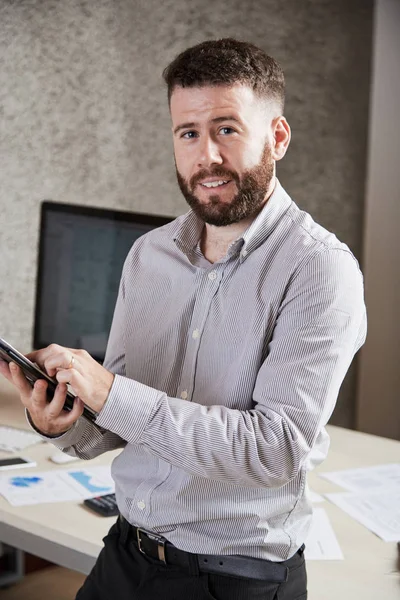 Portrait of bearded office worker leaning on the table using digital tablet in his work and smiling at camera at office