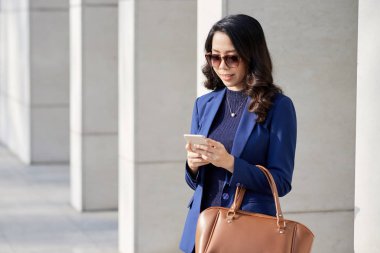 Cep telefonu kullanarak güneş gözlüğü genç iş kadını ve ofis binası nın sütunları açık havada dururken gülümseyerek