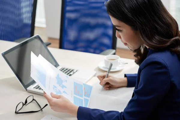 Serious young financial adviser sitting at office desk in front of laptop examining financial charts and making notes in notepad