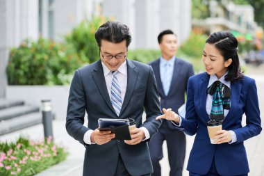 Gülümseyen genç iş kadını kahve içiyor ve o dijital tablet açık havada kullanırken meslektaşı ile online iş tartışıyor