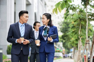Şehirde sokakta yürürken tek kullanımlık bardaklardan kahve içen ve konuşan resmi giyimli iş adamları