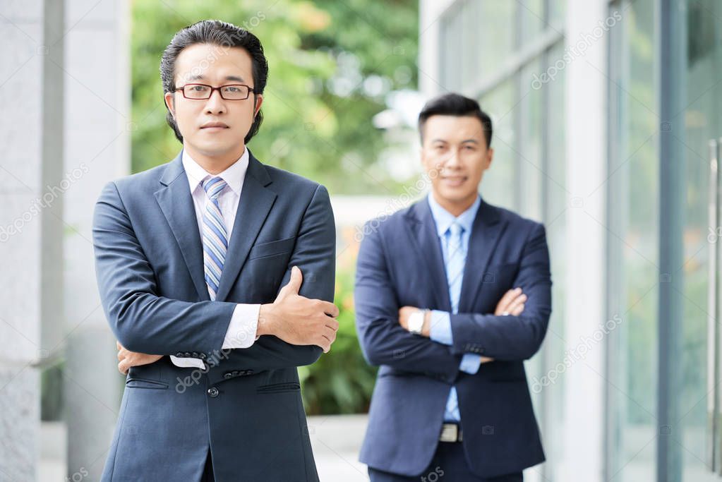 Portrait of Asian two confident businessmen in suits standing with arms crossed and looking at camera outdoors