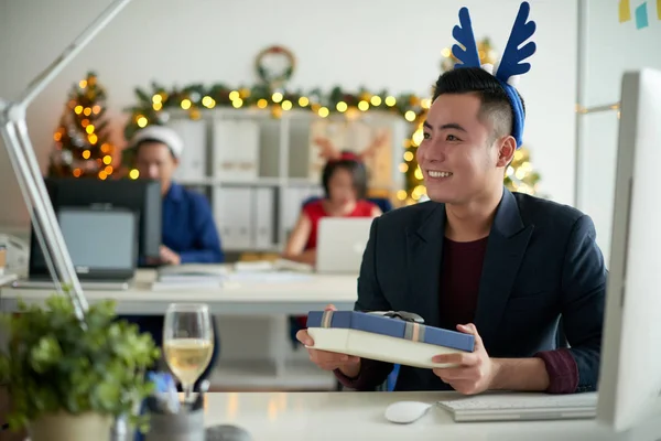 快乐兴奋的年轻亚洲家伙在蓝色鹿角头带坐在桌子上与电脑 并收到圣诞礼物的同事在现代办公室 — 图库照片