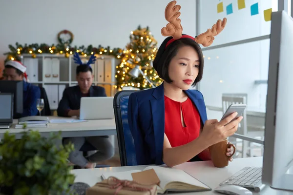 沉思集中的年轻亚洲女士在鹿角头带坐在桌子前的计算机和短信 而在办公室工作 喝咖啡在新年前夕 — 图库照片