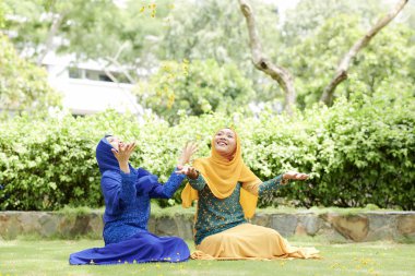 Geleneksel Müslüman elbiseler güzel pozitif genç Vietnamlı kadınlar yeşil çim üzerinde oturan ve yaprakları ile oynarken