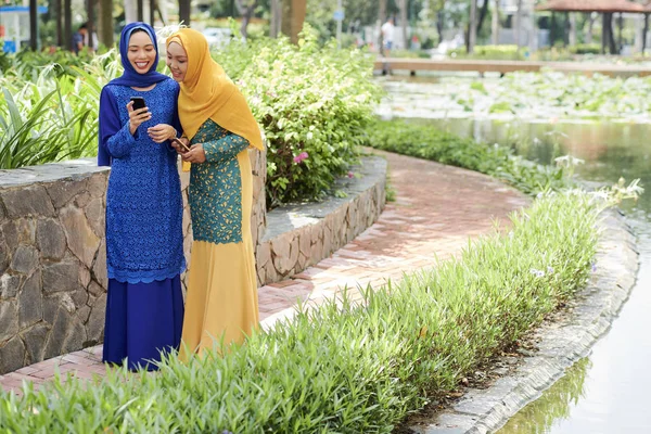 快乐的年轻穆斯林妇女显示短信在智能手机上给她的朋友 当他们走在公园 — 图库照片