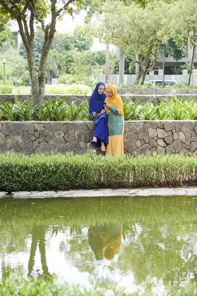 Two Muslim women in hijabs texting friends when spending time in beautiful city park