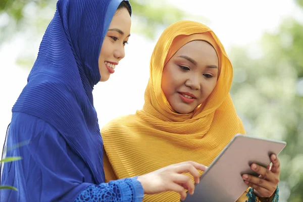 Pretty young Vietnamese woman in blue and yellow hijabs watching something online on digital tablet