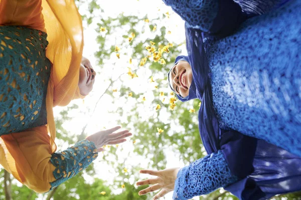 Happy female friends in hijabs throwing flower petals in park, view from below