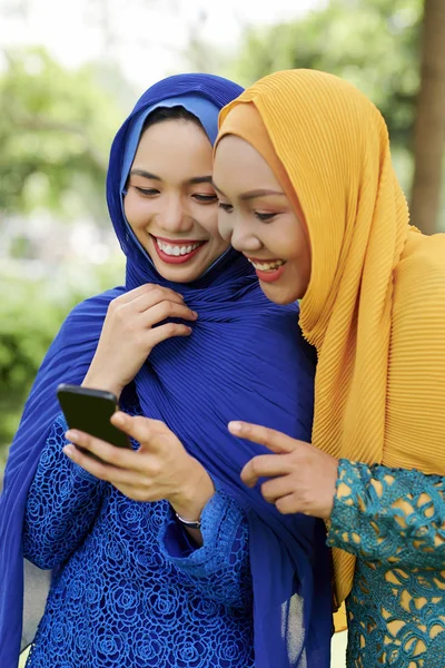 Happy young female friends in hijabs reading text message on smartphone