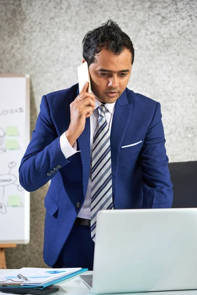 Serious manager in blue suit standing near the table talking on mobile phone and typing on laptop computer at office