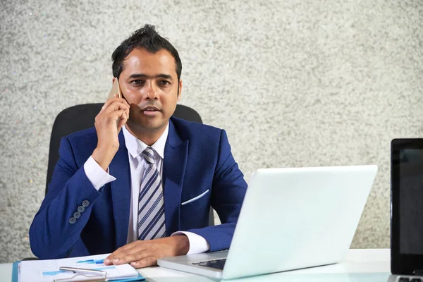 严肃的印度商人坐在笔记本电脑前的桌子前 在办公室用手机交谈 — 图库照片