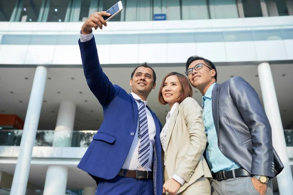 笑顔のビジネスマンは 彼らが屋外のオフィスビルの近くに立っている間 携帯電話で彼の同僚と一緒に自分撮りポートレートを作ります — ストック写真