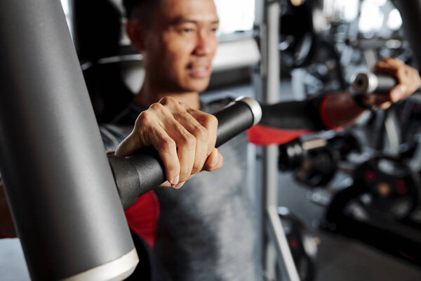 Young positive Vietnamese man doing exercise in fly machine, focus on his hand