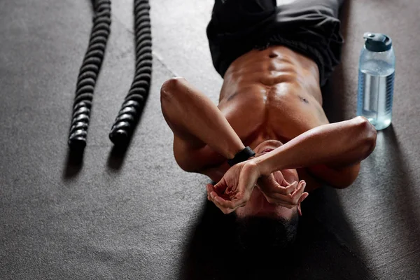 年轻的无衬衫男子躺在地板上 锻炼或休息与战斗绳和瓶水在他附近 — 图库照片
