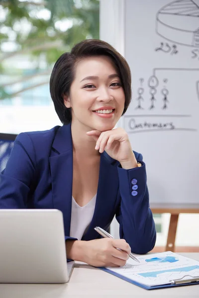 亚洲女高管坐在办公桌前 手提电脑前拿着金融商业计划书 在办公室对着相机微笑 — 图库照片