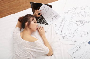 Genç kadın dizüstü bilgisayar ve giysi moda skeçler ile yatakta sıkı çalışma dan sonra uyku