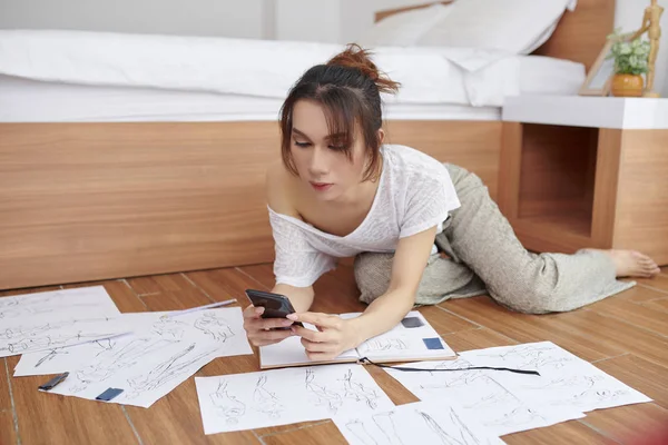Young female designer drawing sketches of clothes for her new collection she lying on the floor and working online on her mobile phone