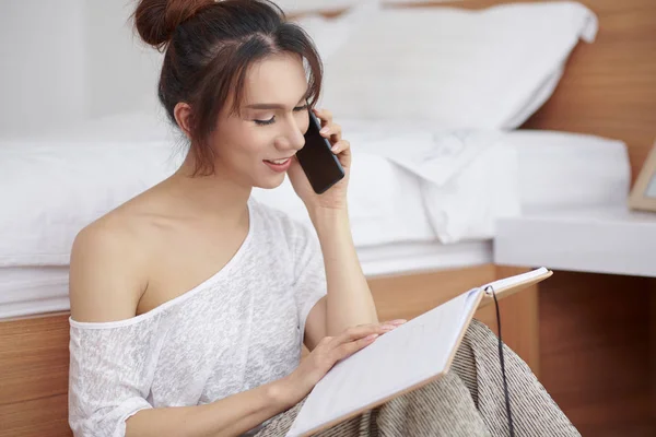 Young businesswoman leaning on the bed while sitting on the floor looking at her notepad and discussing her plans on mobile phone