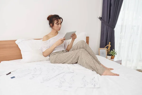 年轻的亚洲妇女坐在她的床上 在她周围画素描 在家里的平板电脑上看的东西 — 图库照片