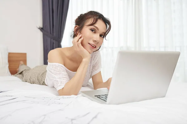 若いデザイナーの肖像画は ラップトップコンピュータで作業し 彼女の寝室でカメラを見てファッションスケッチの間にベッドに横たわって — ストック写真
