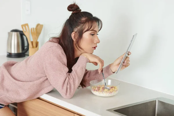 年轻女孩拿着数字平板电脑 看着它的东西 而吃玉米片与牛奶在家庭厨房早上 — 图库照片
