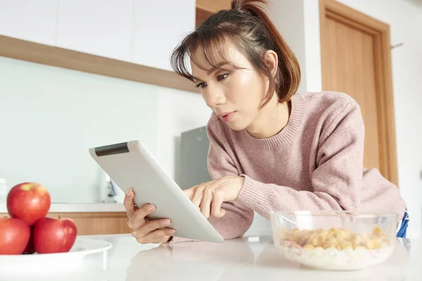 キッチンで朝食中にミルクと新鮮なリンゴとコーンフレークを食べながら オンライン作業のためにデジタルタブレットを使用してアジアの若い女の子 — ストック写真