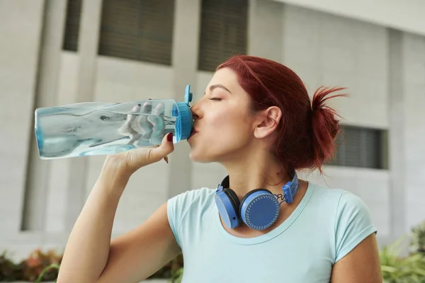 年轻美丽的健康女人与耳机在她的脖子上喝淡水 — 图库照片