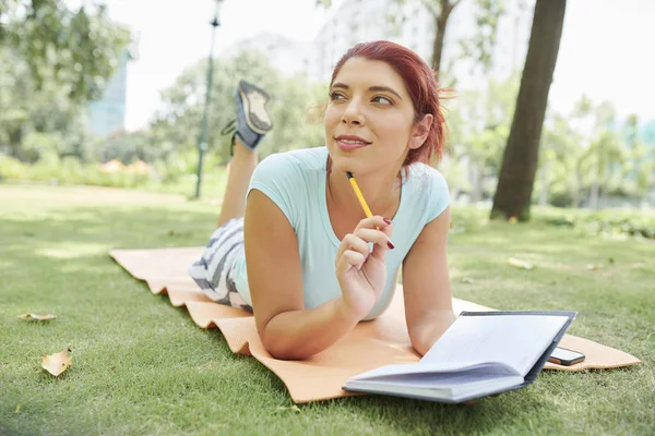 Pensive pretty young woman lying on yoga mat in park and writing ideas in her planner