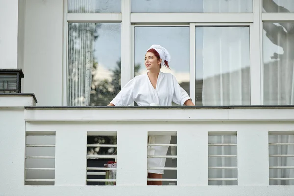 Attractive smiling young woman in bathrobe standing at balcony and enjoying beautiful morning