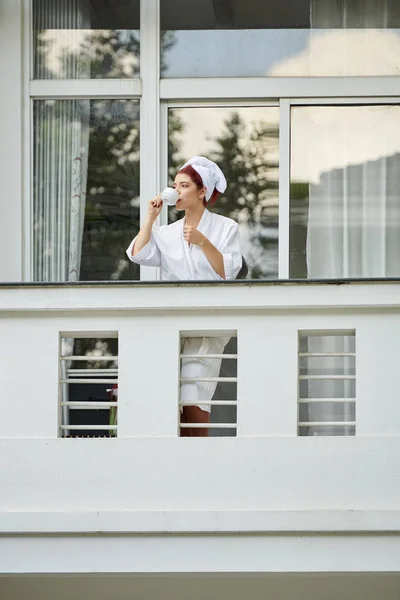 美丽的年轻女子穿着浴袍站在阳台上 喝着早晨的咖啡 — 图库照片