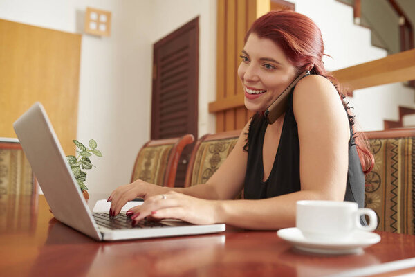 Cheerful beautiful business lady answering e-mails on laptop and talking on phone