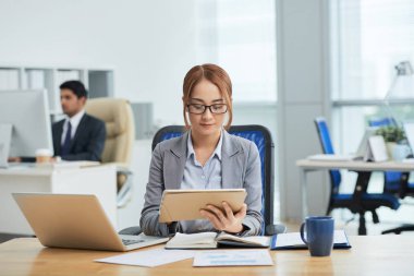 Gözlüklü genç iş kadını işyerinde oturan ve işine konsantre o arka planda çalışan işadamı ile online iş için dijital tablet kullanarak