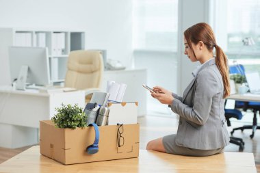 Genç Asyalı çalışan ofis malzemeleri kutusu ile masada oturan ve cep telefonu kullanarak, o yeni bir iş almak