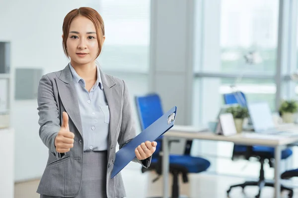 Portrait of young Asian businesswoman standing at office with folder looking at camera and showing thumb up