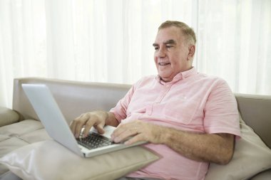 Mutlu kıdemli adam kucağında yastık ile kanepede oturan ve dizüstü bilgisayar üzerinde çalışıyor