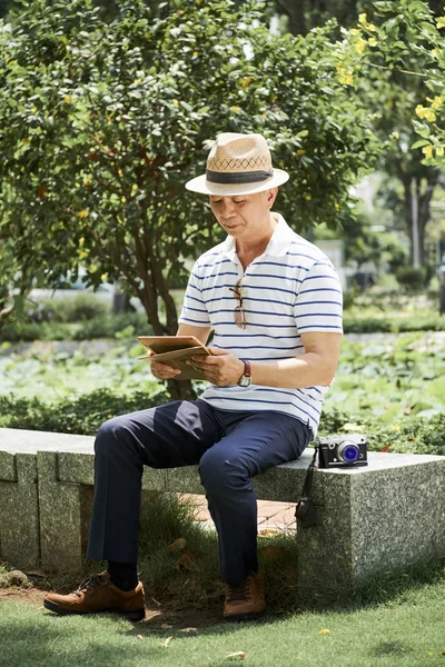 공원에서 카메라와 벤치에 앉아있는 디지털 태블릿을 사용하여 캐주얼 웨어와 모자에 — 스톡 사진