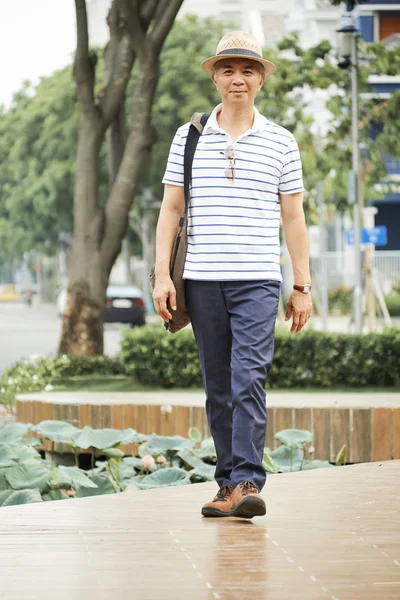 Şapkalı ve gündelik giyimli Asyalı yaşlı adamın portresi parkta yürürken ve yaz günü kameraya bakarken
