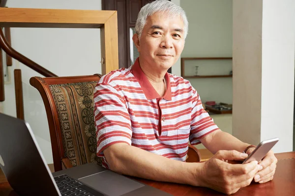 亚洲自信男子的肖像 使用他的手机和在线交流 而坐在桌子上与笔记本电脑 看着相机 — 图库照片