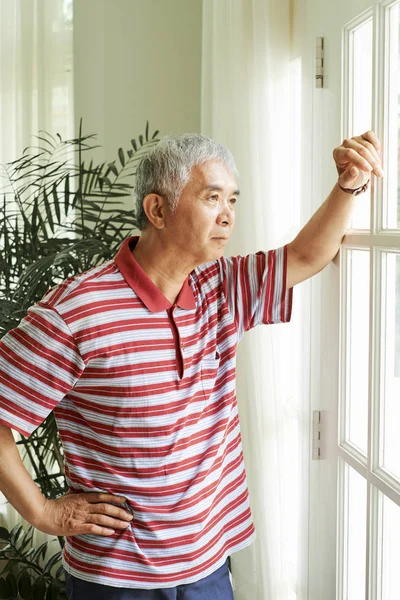 Pensive Asian man standing near the glass door looking at view or waiting for somebody