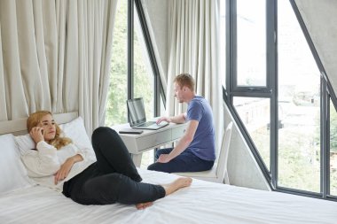 Kız arkadaşı yatakta dinlenirken ve telefonda konuşurken dizüstü bilgisayar üzerinde çalışan genç adam